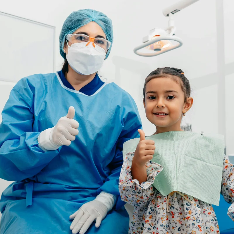 Pediatric dentist in blue scrubs, mask, and gloves giving thumbs up with a smiling young girl patient in bib during child-friendly dental check-up at Ridansh Hospital.